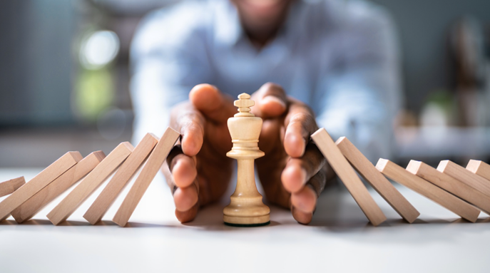 Hands protecting a chess king from falling dominoes, representing risk prevention and proactive safety